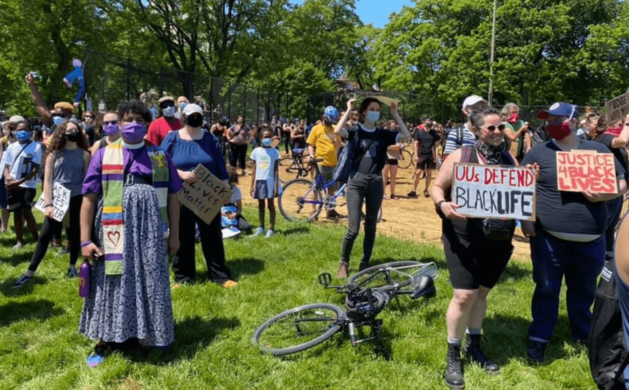 A large group gathers outdoors, many wearing masks, some holding various 