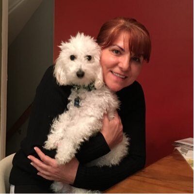 a woman with red hair pulled back and bangs wears a black shirt and holds a fluffy white dog